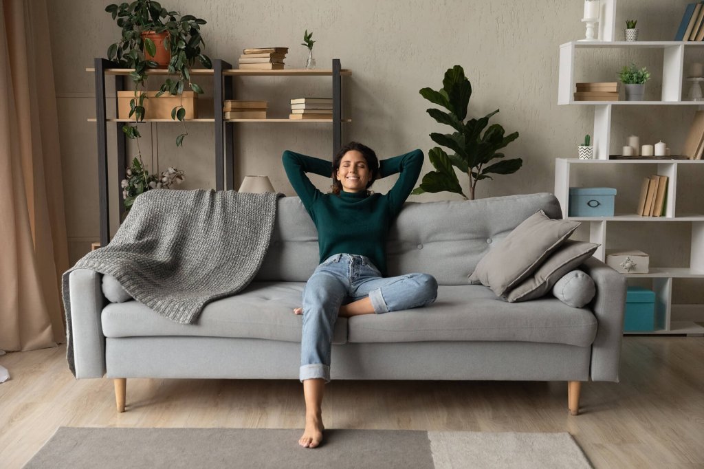 A lady is sat in the centre of a living room on a sofa with her eyes closed, her arms behind her head and with one leg up on the sofa. In the background, there are white and black shelved cabinets with plants and books placed on them. In the foreground, there is a large rug.