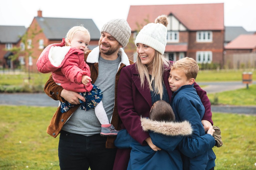 A family are standing together on a grass area in front of a newly built estate. The father is holding their daughter and their two sons are hugging their mother. They are all dressed in winter clothing.