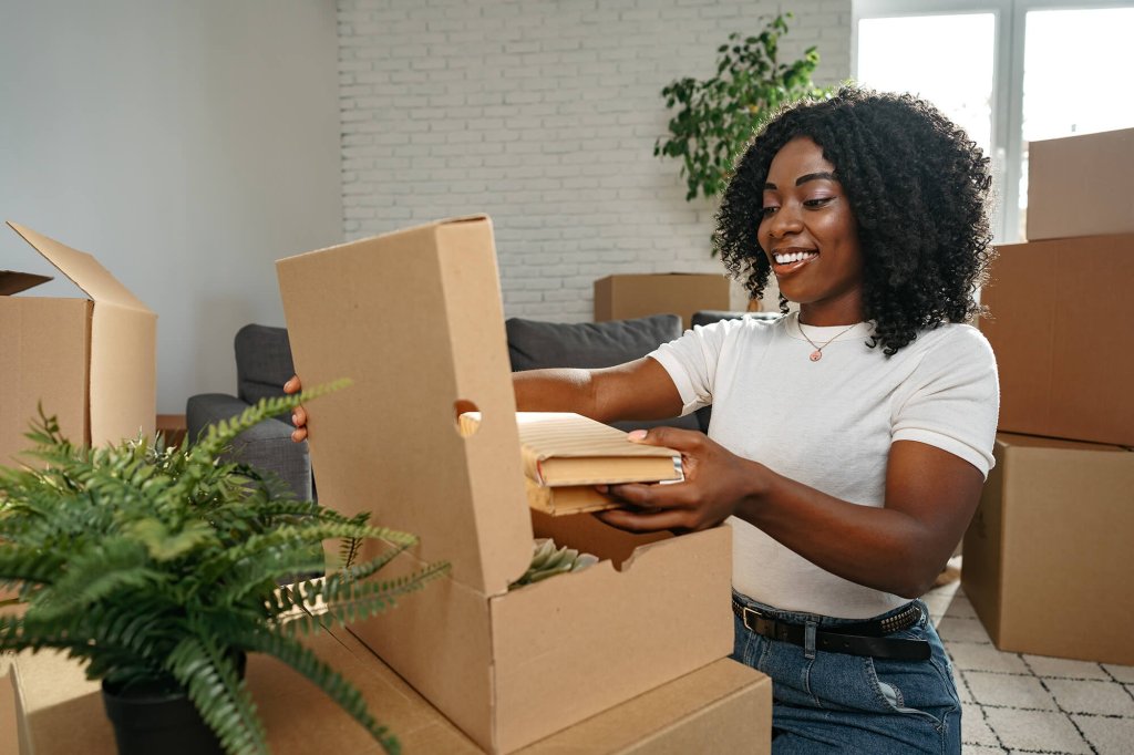 A woman with curly hair is opening a cardboard box and removing house hold items to put into a new home living space. She is surrounded by stacked boxes and there is a plant in the foreground resting on one of the boxes.
