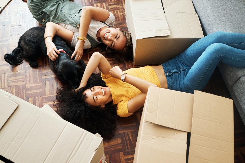 Two females and a dog lay on the floor of a new home surrounded by moving house boxes. One of the woman has her legs up on a sofa and they are both laughing.