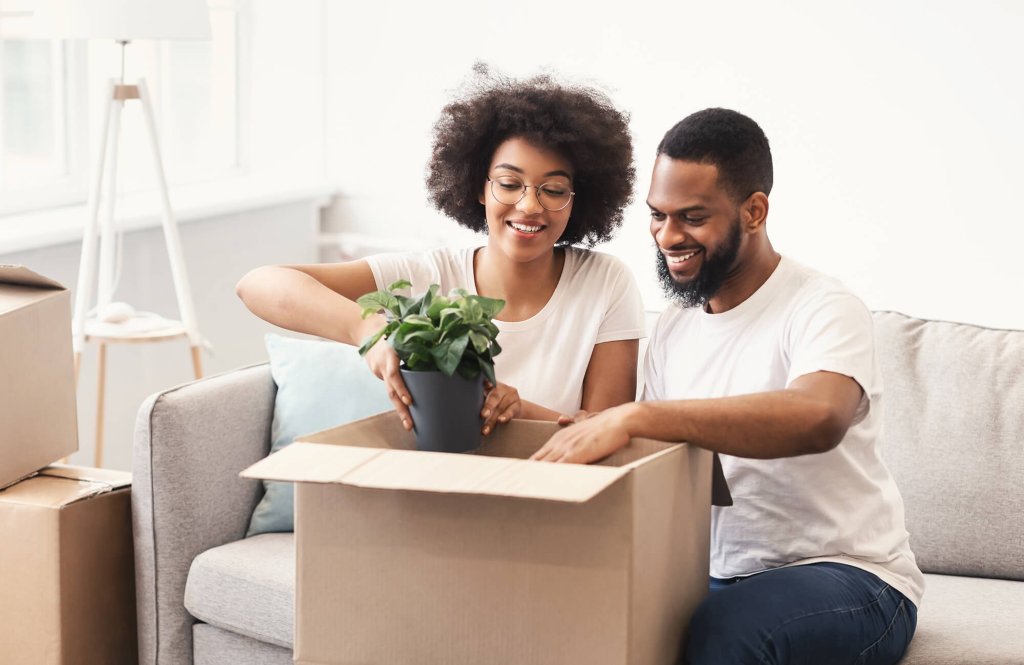 A couple sitting on a sofa packing household items into moving house boxes. The lady is placing a plant into the box while the man helps by holding the box open.
