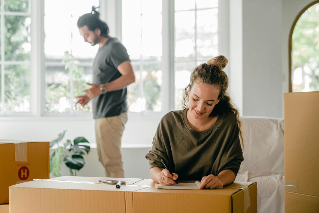 A woman in writing a note on top of a sealed moving box in a living room space. There is a male standing in the background near other sealed boxes.