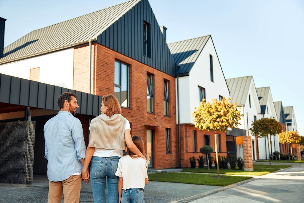 A couple are standing outside a new home with their young daughter in a suburban neighbourhood.