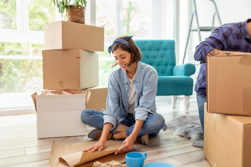 A woman sitting on the floor packing house items into moving boxes, surrounded by moving supplies, more boxes and an armchair in the background. There is a males arm to her left who is sealing the boxes shut.