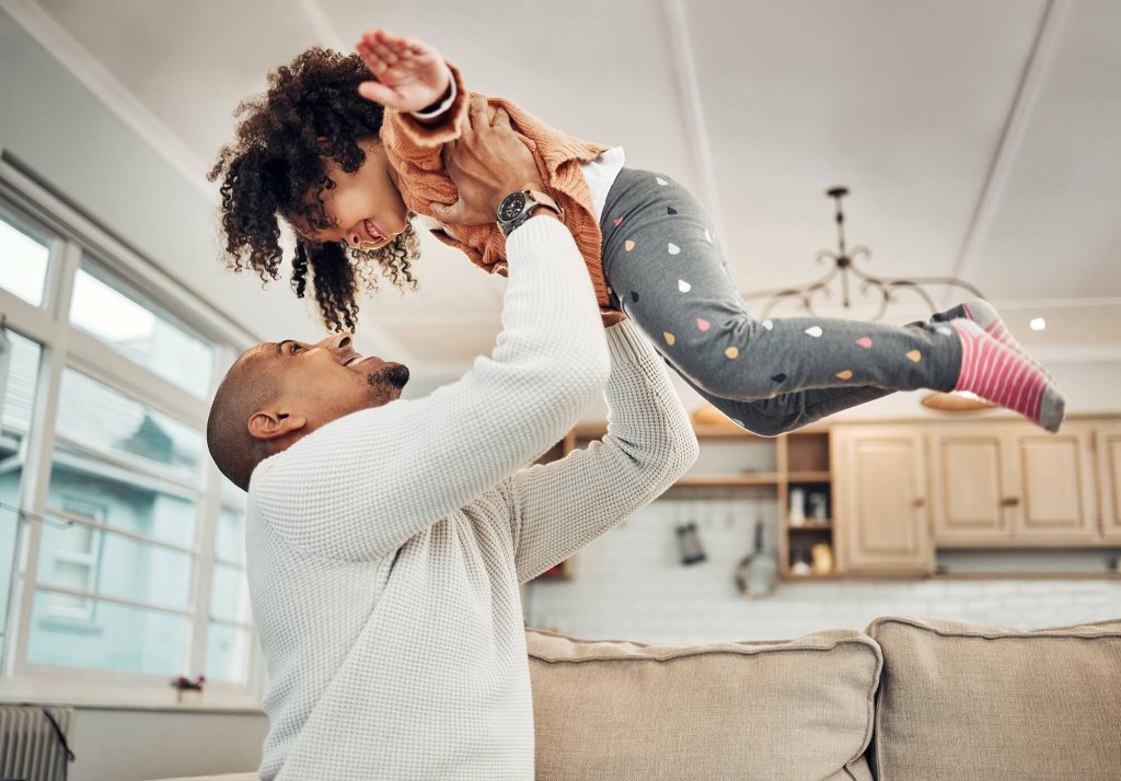 A father is lifting his young daughter in the living room of their home. Her arms are out in an aeroplane motion and they are both smiling.