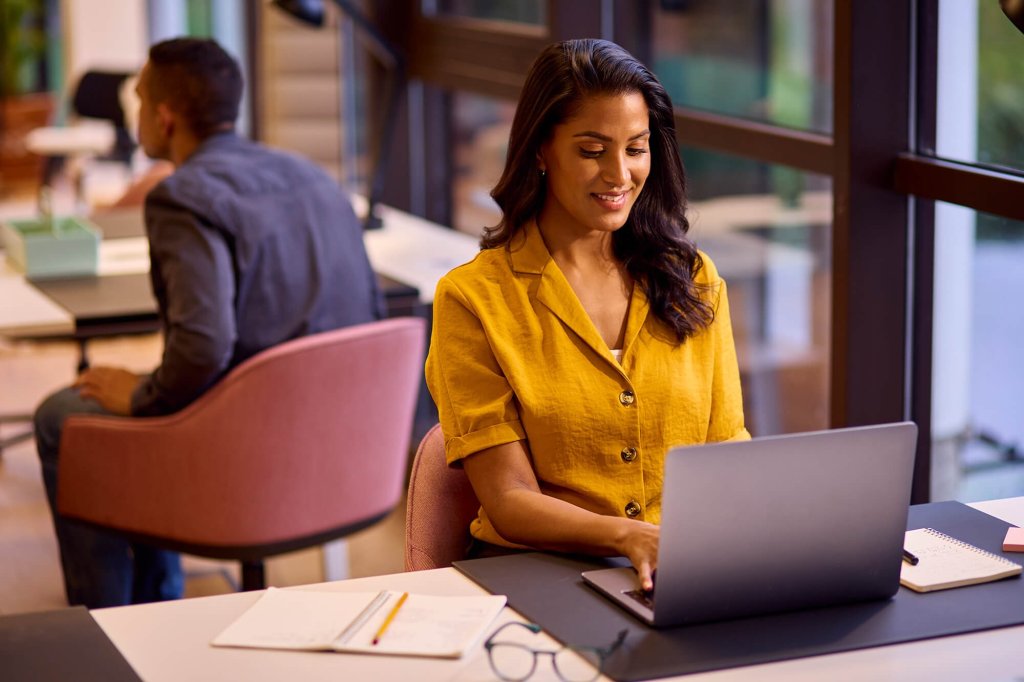 A female solicitor in a buttoned blouse is sat working on a laptop at her desk in an office. There is a notebook, pen and reading glasses on her office desk. A male solicitor is sitting on a chair at another desk in the background.