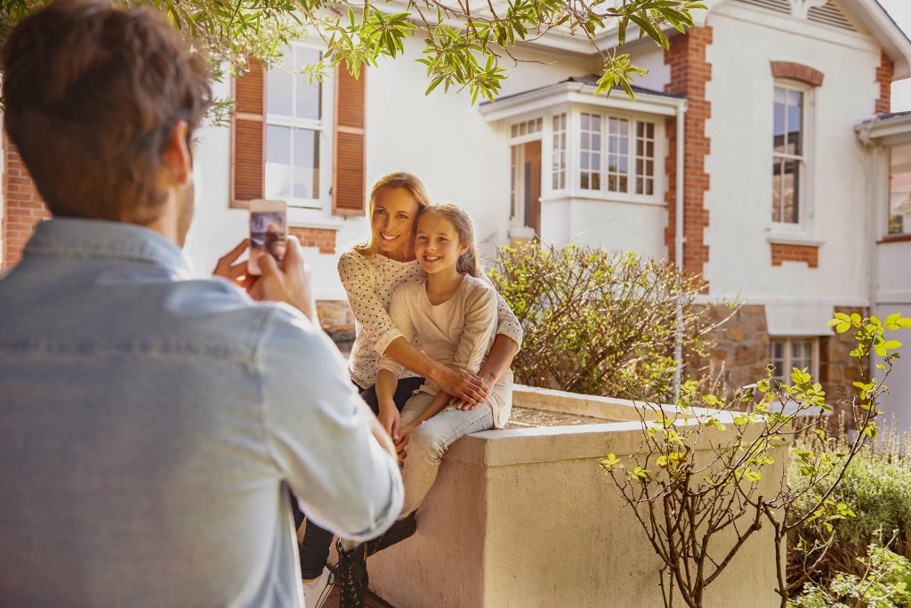 A man is taking a photo of a woman and a young girl who are smiling. The woman has her arms around the girl and they are both sitting on a stone wall in front of a house.
