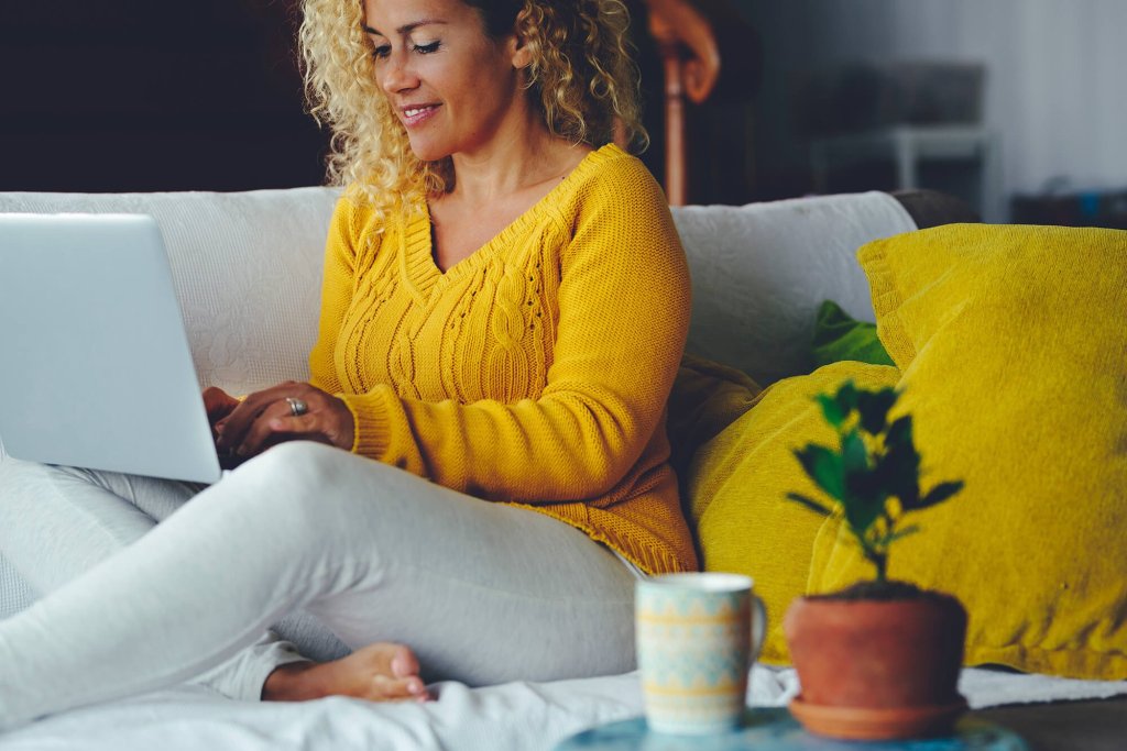 A woman with curly hair, wearing a sweater, sitting on a sofa in a living room using a laptop to search for conveyancing quotes. A potted plant and a mug are on a table in the foreground.