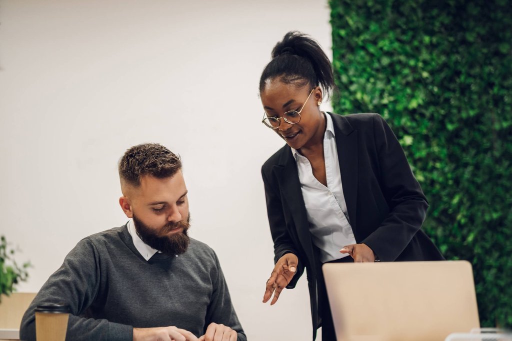 A lady is stood up in an office space talking a man, who is sitting at a desk, through solicitor's documents. They are both wearing smart clothing and there is a takeaway coffee cup and laptop on the desk. The office around then is a bright space with a decorative leaf wall.