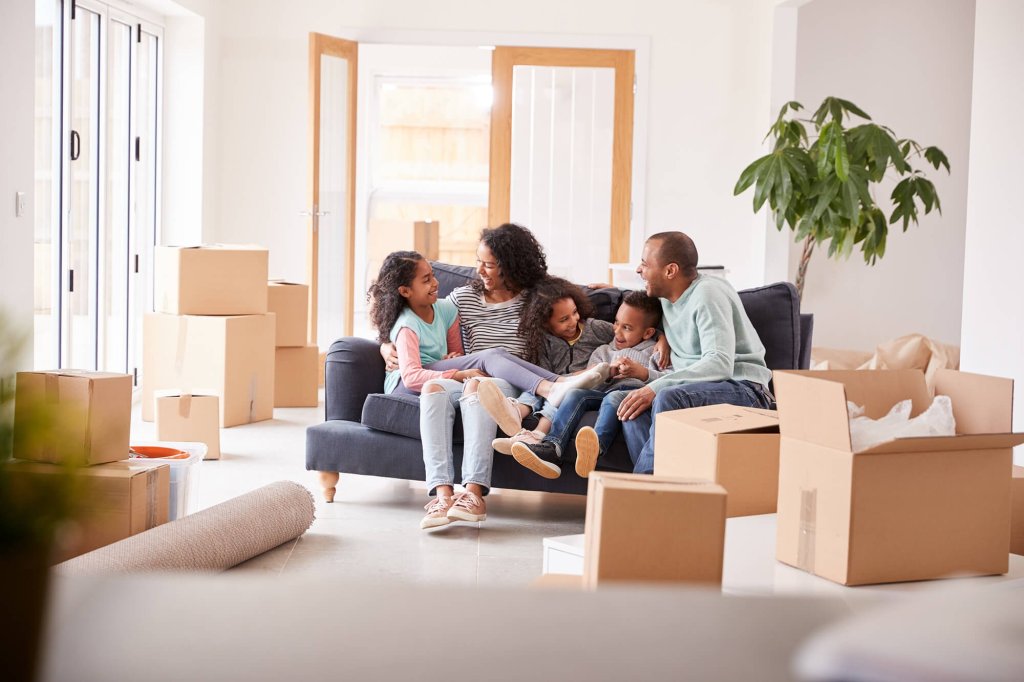 A family are sat together on a sofa in the living room of a new home surrounded by moving boxes. The three young children, mother and father are all happy.