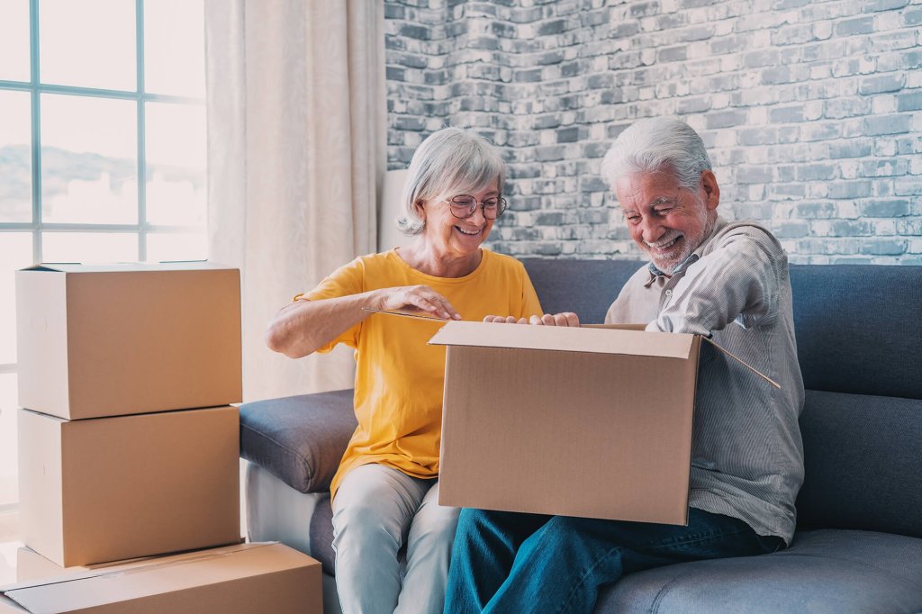 An elderly man and woman are sitting on a sofa unpacking boxes in the living room of their new home. There is a stack of unopened boxes next to the woman. They are both smiling and are dressed in casual clothing.
