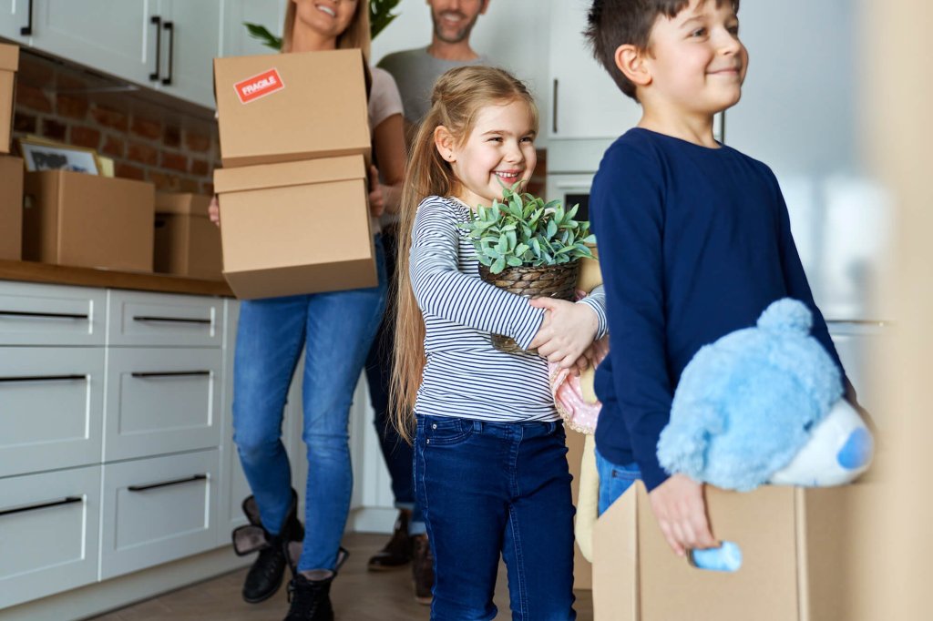A family that are selling their home are exiting the kitchen with packed boxes. The mother, father and little boy all smile as they carry a box out of the kitchen. The little girl is holding a plant and is walking behind the little boy. There are multiple boxes on the kitchen countertop.