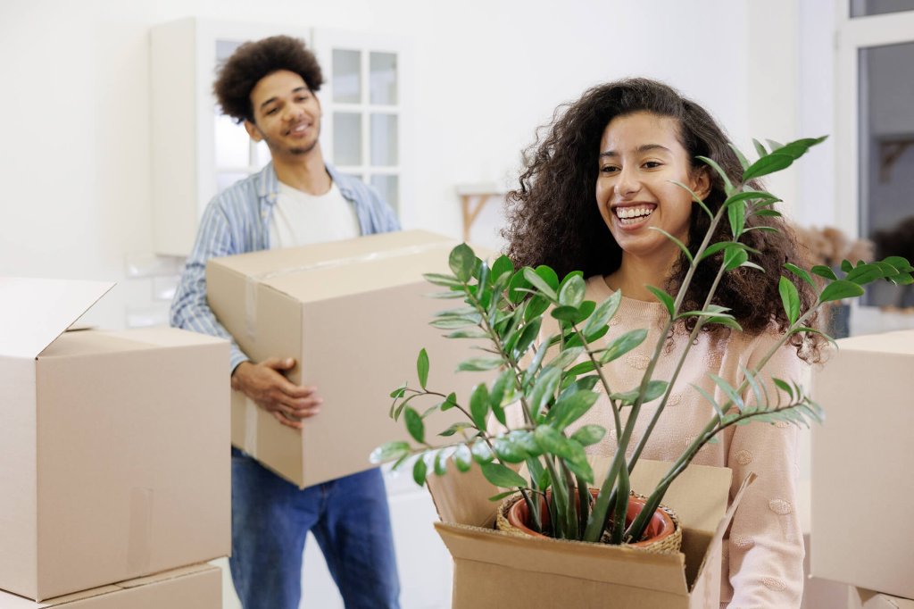 A young male and a young female are smiling while carrying boxes in a new home. The female is carrying an open box with a plant inside of it while the male carries a box that is sealed shut.